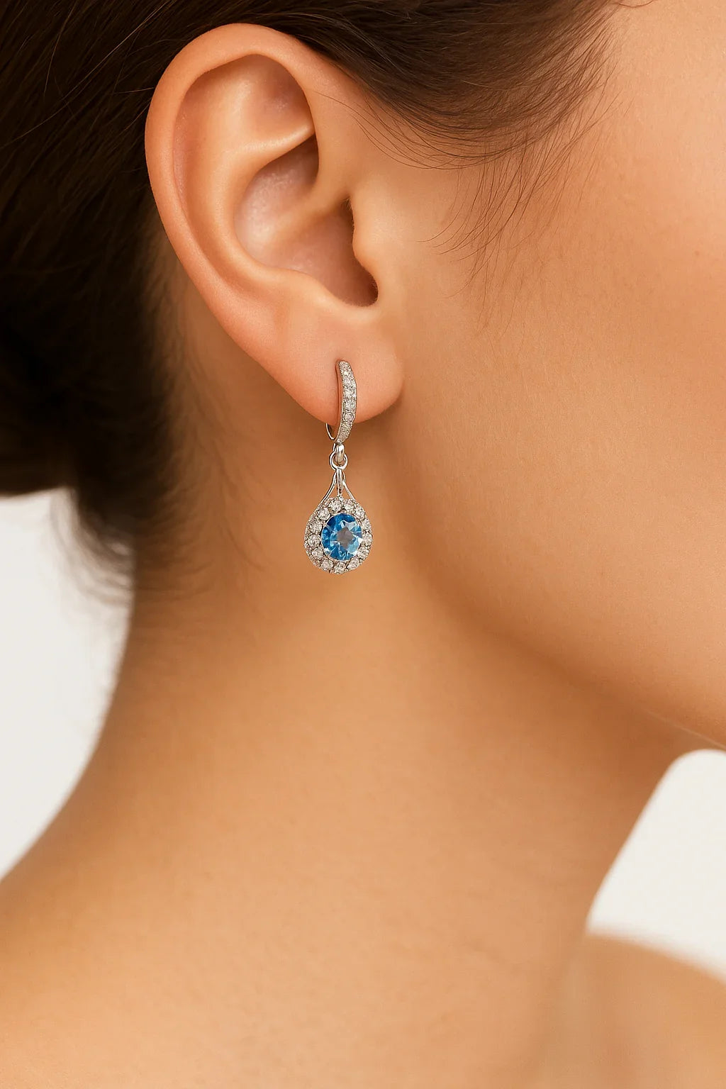 Close-up of woman's ear wearing silver drop earrings with blue moissanite and surrounding white stones