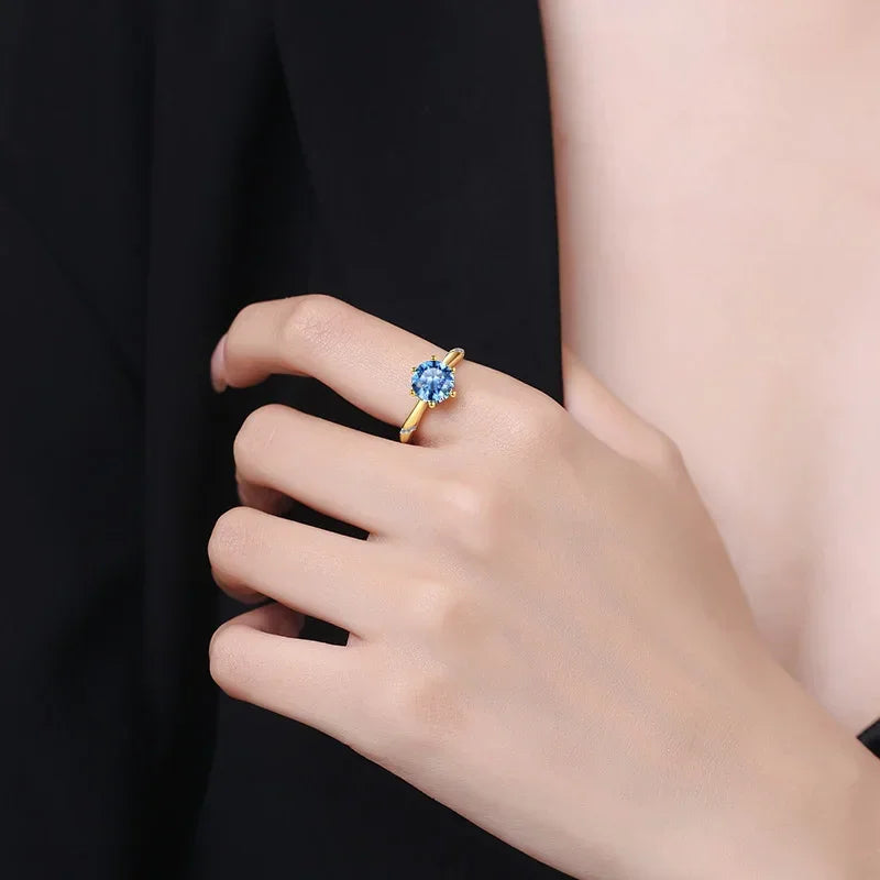Hand wearing 18k gold ring with a 2ct round blue moissanite stone against black background